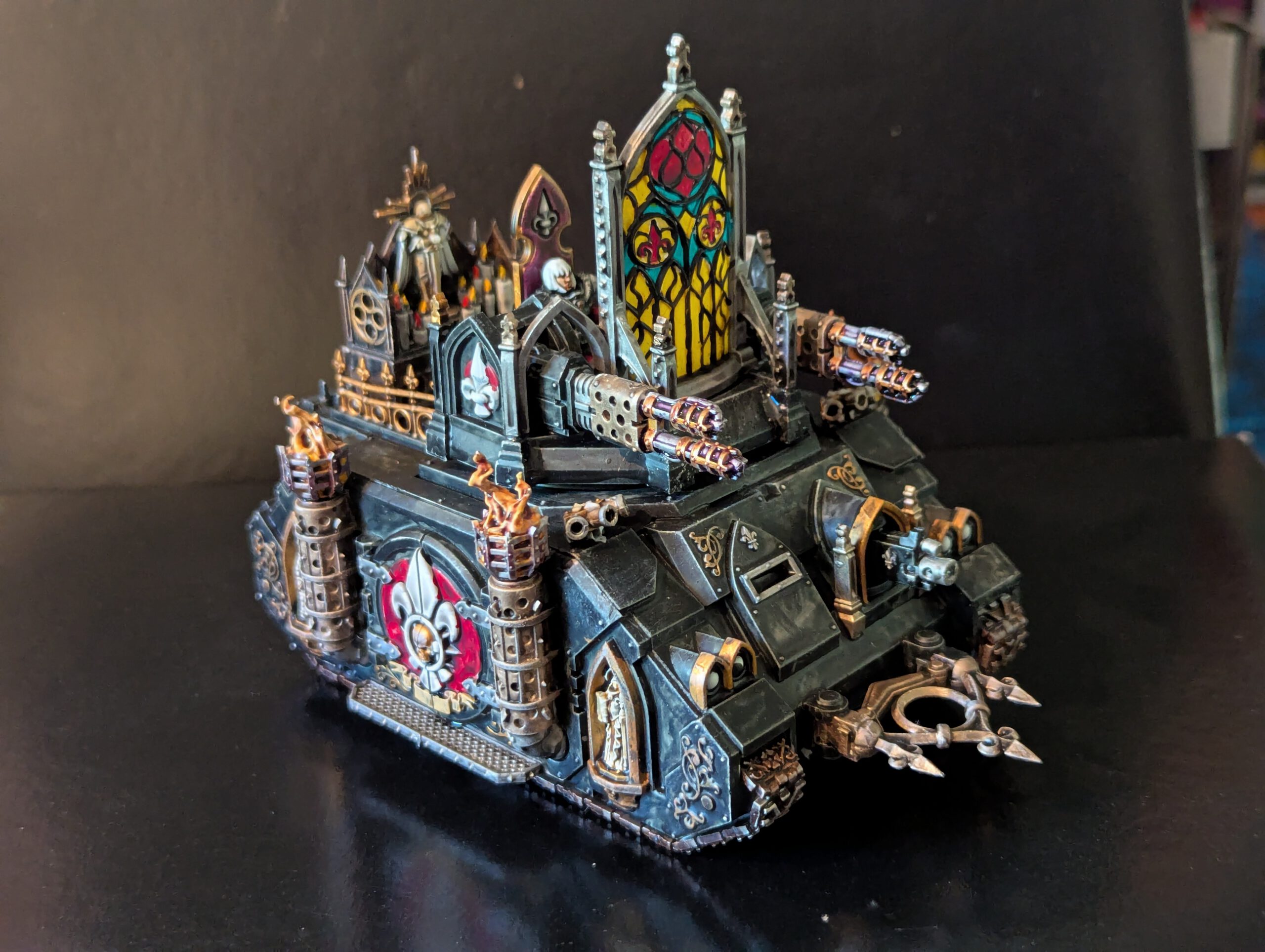 A fully painted Adepta Sororitas Immolator tank in black armor with gold trim, featuring a vibrant yellow and red stained-glass sanctuary and twin-linked heavy bolters.